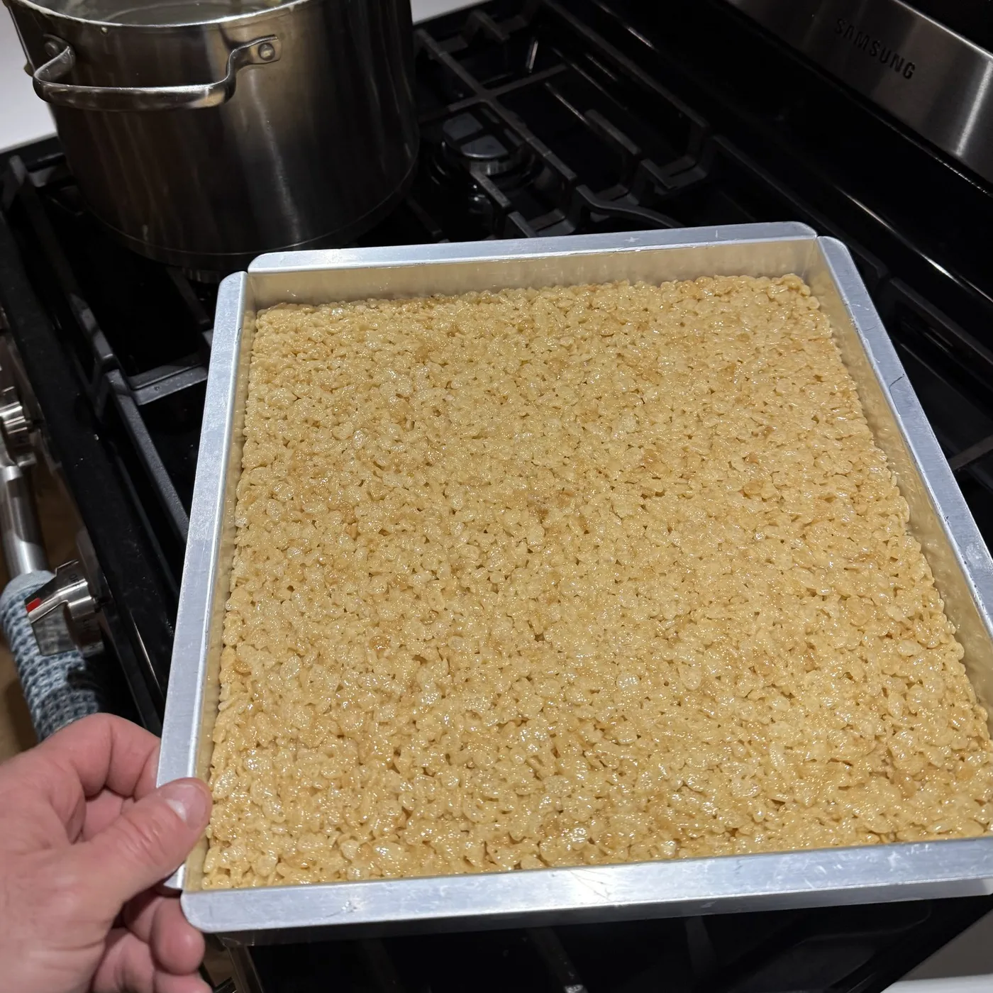 A 12x12 aluminum pan packed flat with krispy brick mix, held over the stovetop beside the pot the butter and marshmallows melted in.