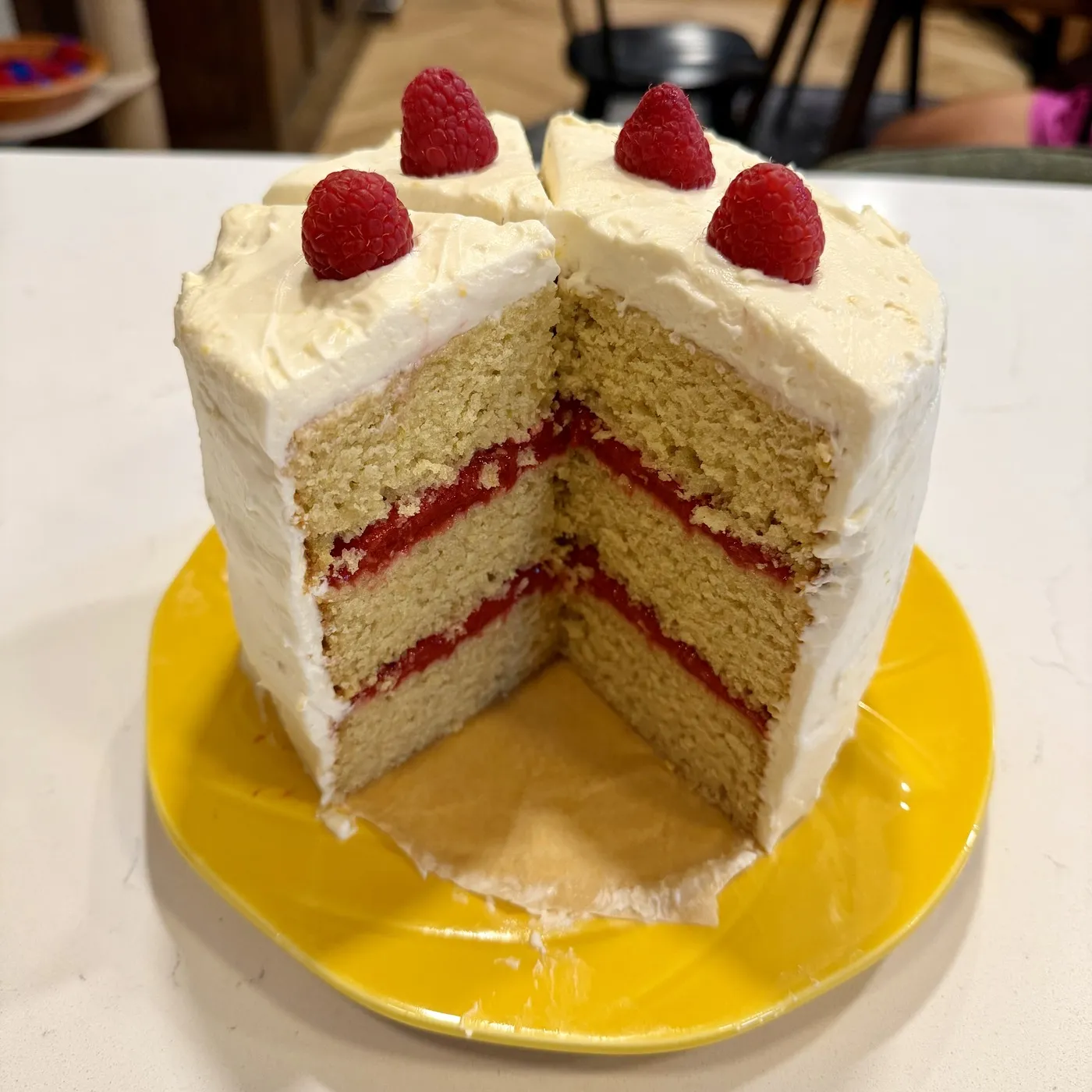 The cake cut open on a yellow plate, three sourdough layers with raspberry between them under white lemon cream, four raspberries on top.