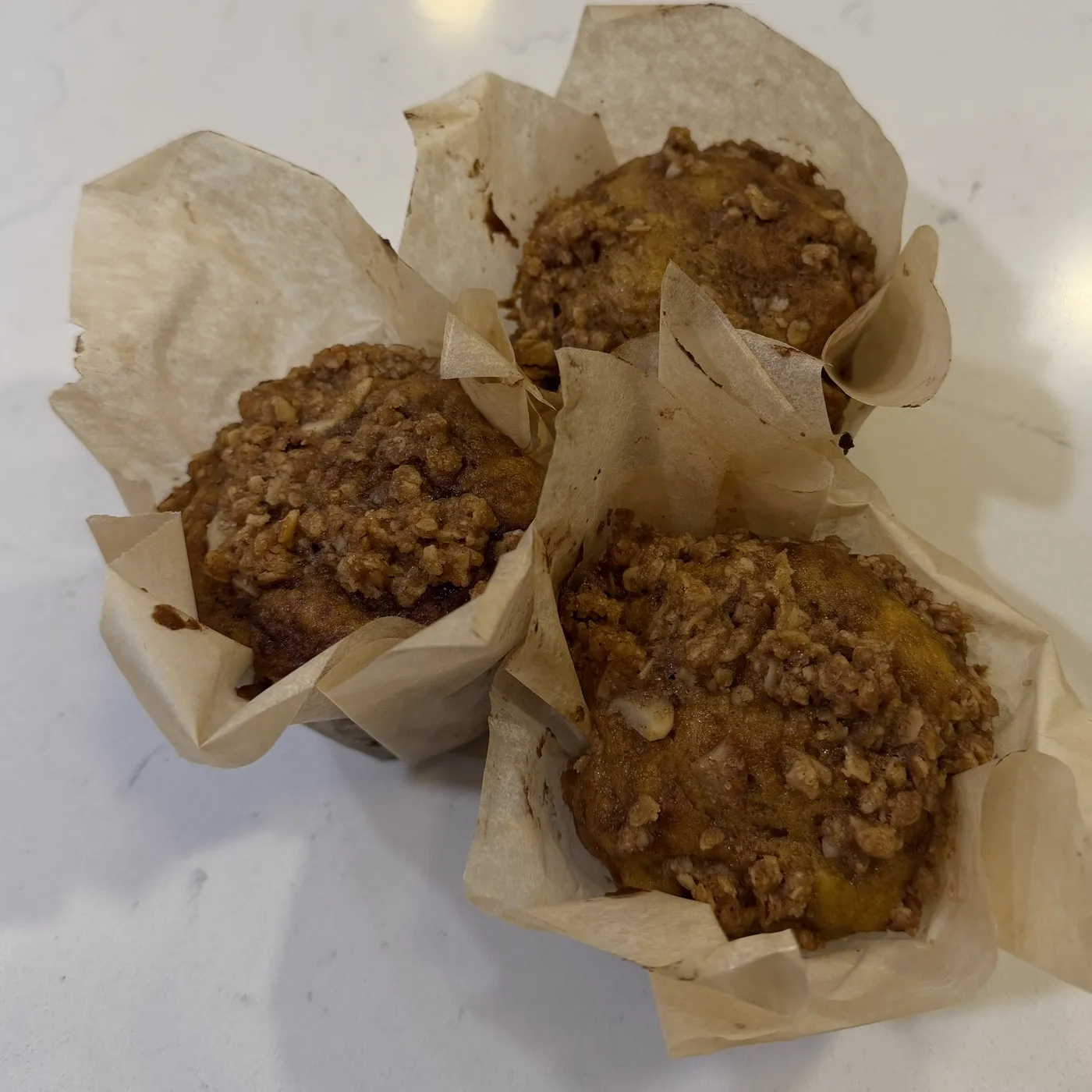 Three pumpkin banana muffins in parchment wrappers on a counter, oat and almond crumble across the tops.